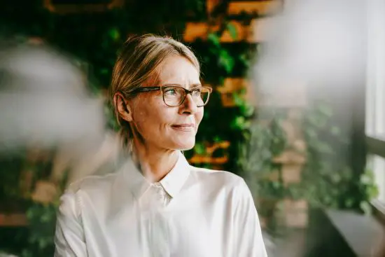 Kvinna med glasögon och vit skjorta ser ut genom fönster, omgiven av gröna växter