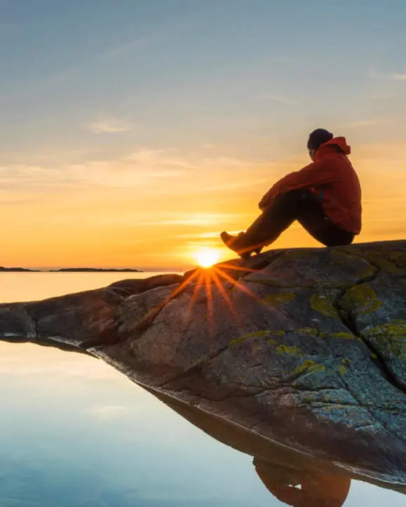 Person sitter på klippa och tittar på solnedgång över havet, med klar himmel och reflektion i vattnet
