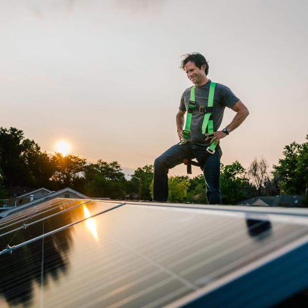 Man installerar solpaneler på taket vid solnedgången, utrustad med säkerhetssele, i en naturskön omgivning