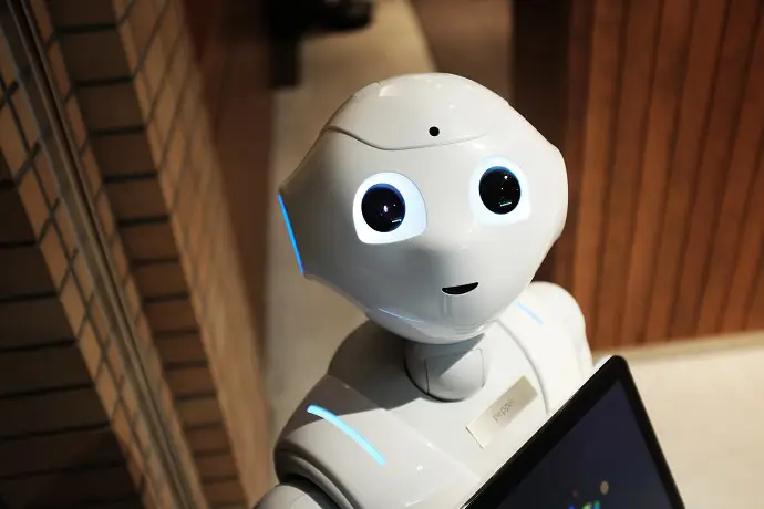 Små leende humanoid robot som står inomhus med en skärm i händerna, omgiven av trädetaljer