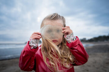 Flicka i röd regnjacka på strand håller något transparent framför ansiktet