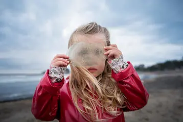 Flicka i röd regnjacka på strand håller något transparent framför ansiktet