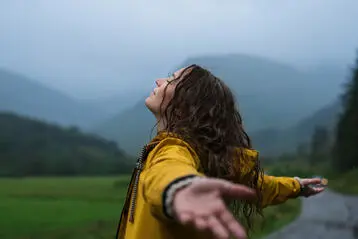 Kvinna i gul regnjacka sträcker ut armarna i en dimmig bergslandskap