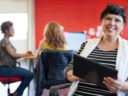 Leende kvinna med kort hår håller i en surfplatta på ett modernt kontor, med kollegor i bakgrunden som jobbar vid datorer