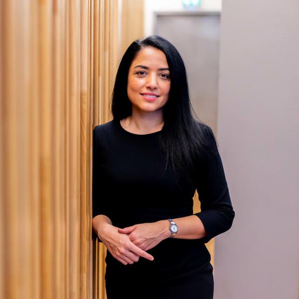 Kvinna i svart klänning står lutad mot en träpanelvägg och ler