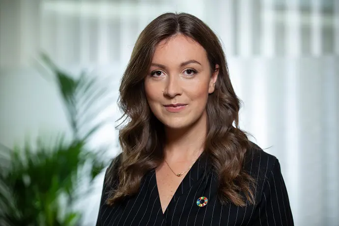 Kvinna med brunt hår i svart randig kavaj, står framför en suddig bakgrund med grönska. Hon bär en pin med färgglada cirklar