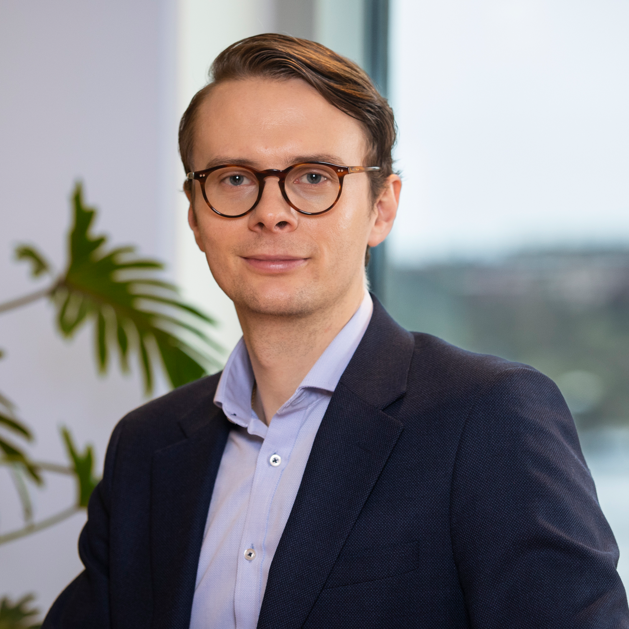 Man i kostym med glasögon sitter i ett kontor framför en växt och ett fönster