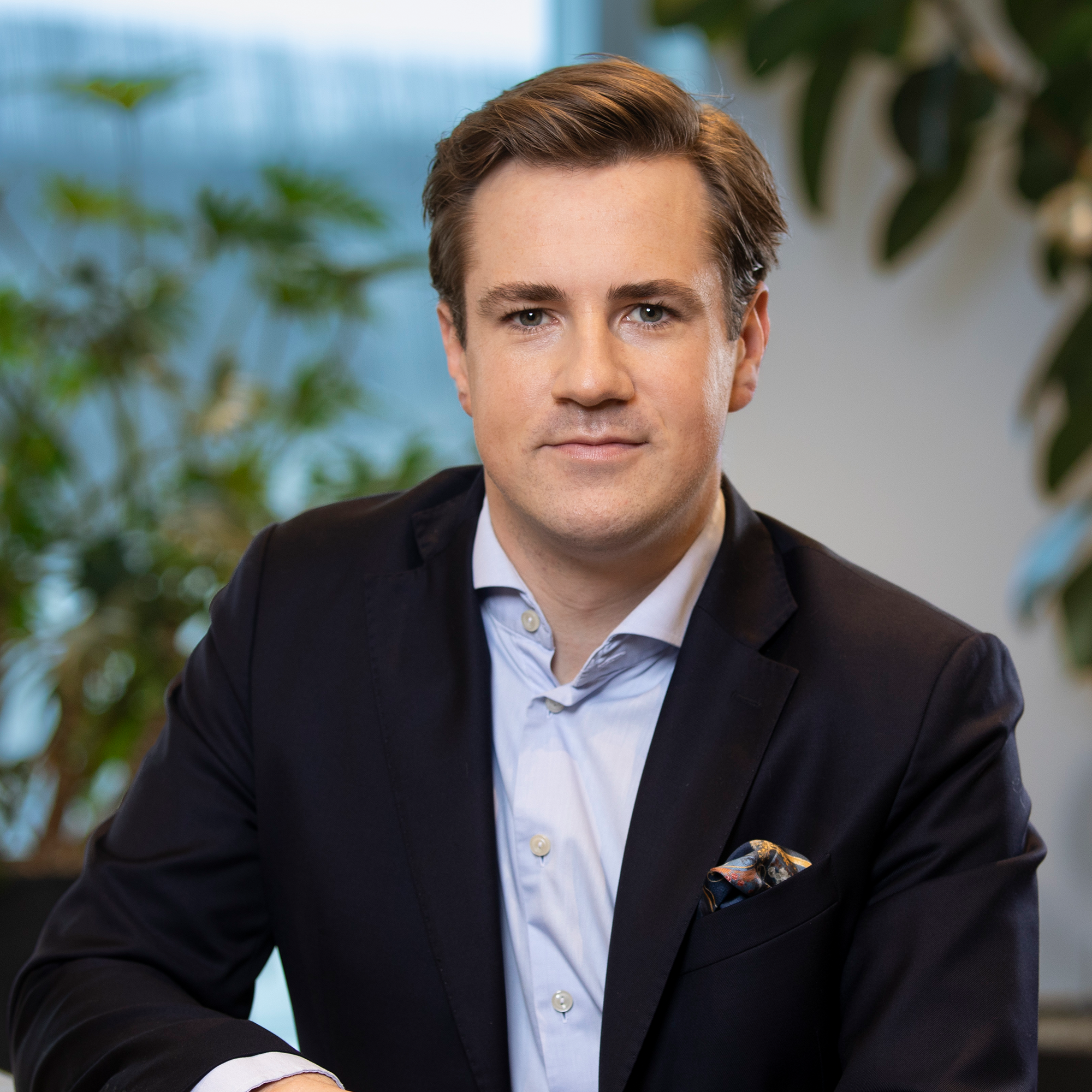 En man i kostym sitter inomhus med växtlig bakgrund