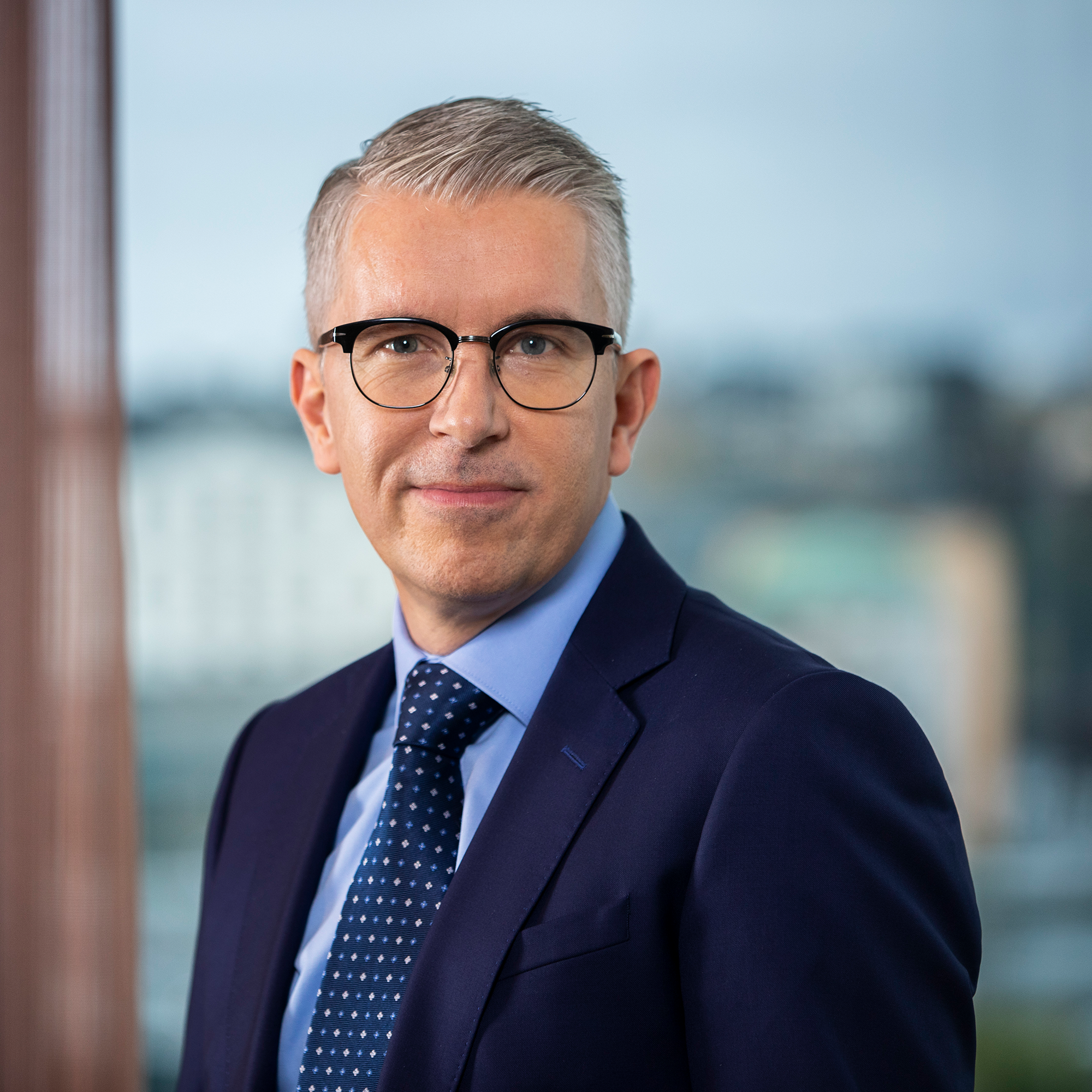 En man i kostym med glasögon och grått hår står framför en suddig bakgrund av byggnader