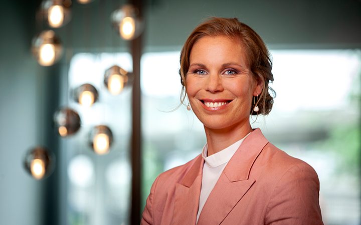 Kvinna i rosa kavaj ler mot kameran i mjukt belyst inomhusmiljö