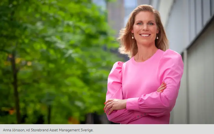 Kvinna i rosa klänning står utomhus med korsade armar och ler, med grönska i bakgrunden och en kontorsbyggnad till höger