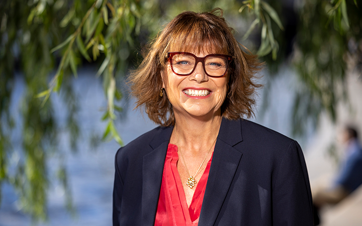 Kvinna med kort hår och glasögon ler framför en suddig bakgrund av grönska och vatten. Hon bär en mörk kavaj och en röd blus