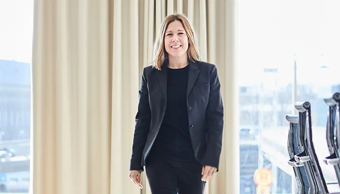 Kvinna i svart kostym står leende framför ett fönster med gardiner i kontorsmiljö