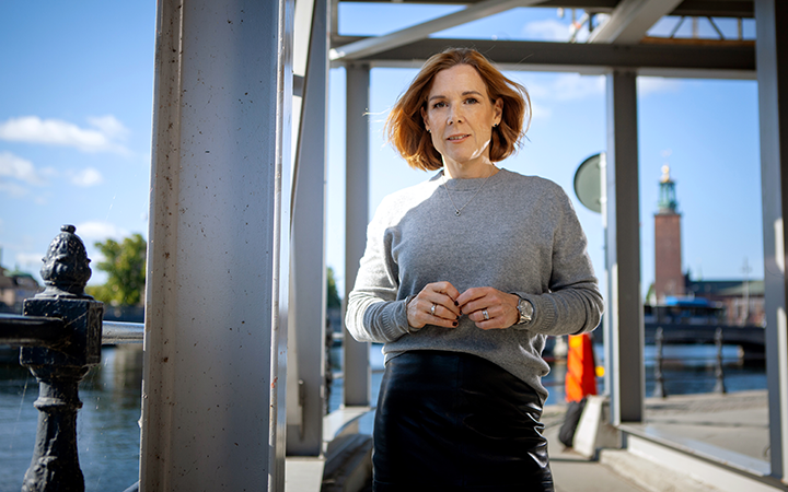 Åsa Wallenberg i grå tröja står vid vattnet i Stockholm med Stadshuset i bakgrunden