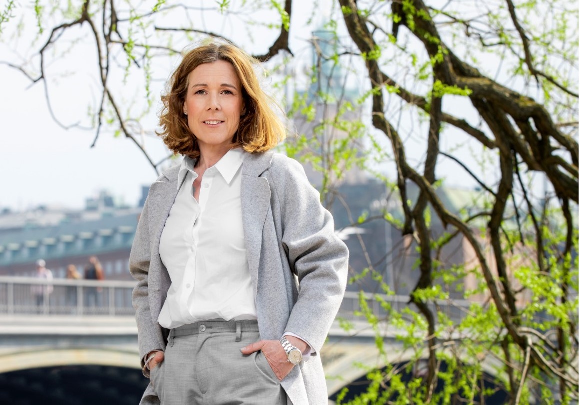 Kvinna i grå kavaj står framför en bro i Stockholm, omgiven av grönskande träd, under en klar vårdag