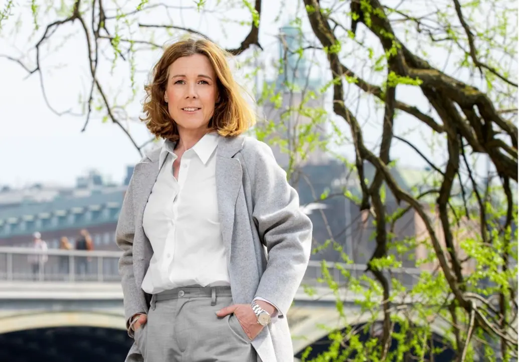 Kvinna i grå kavaj står framför en bro i Stockholm, omgiven av grönskande träd, under en klar vårdag