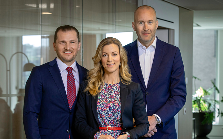 Tre affärsmän i kostymer står tillsammans på ett modernt kontor, leende mot kameran