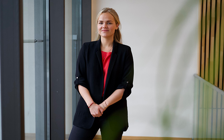 Kvinna i svart kavaj och röd topp står vid ett fönster och ler, inomhusmiljö