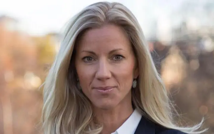 Kvinna med blont hår i affärsmässig klädsel står utomhus med en suddig bakgrund av träd i höstfärger