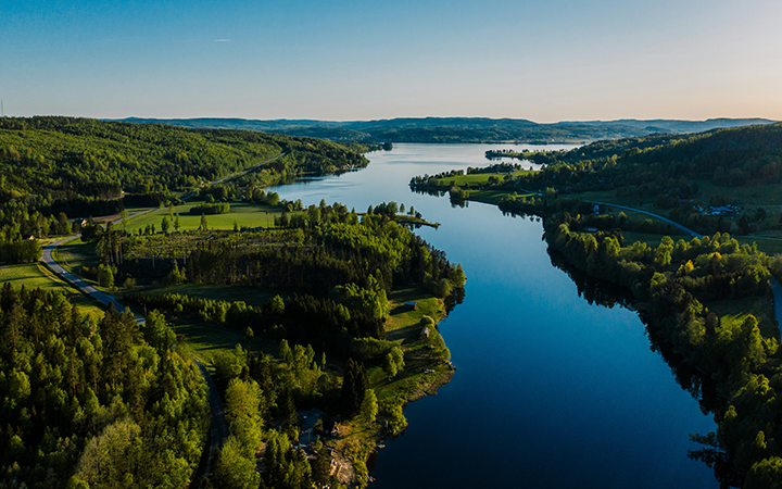 Flygbild över en lugn sjö omgiven av frodiga skogar och gröna landskap i Sverige, under solig himmel
