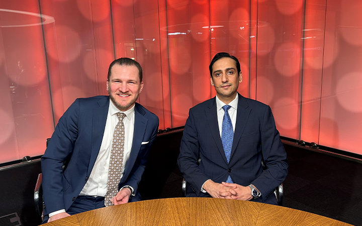 Två affärsmän i kostymer sitter vid ett bord framför en röd bakgrund