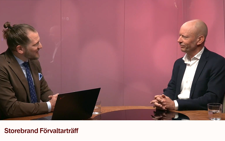 Två män diskuterar vid Storebrands förvaltarmöte, sittande vid ett bord med en bärbar dator och glas på en rosa bakgrund