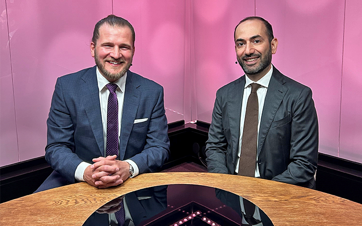 Två affärsmän i kostymer sitter vid ett runt bord mot en rosa bakgrund