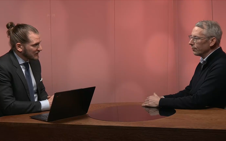 Två män i kostymer sitter vid ett träbord och samtalar, en med laptop, mot en röd bakgrund i en kontorsmiljö