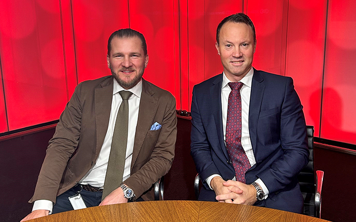 Två affärsmän i kostym sitter vid ett bord med en röd bakgrund