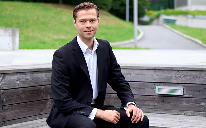 Geir Magne Boei svart kostym sitter utomhus på en träbänk, omgiven av grönska och en asfalterad gångväg