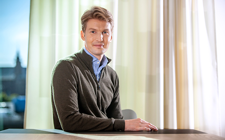 Man i mörk tröja som sitter vid ett bord med ljusa gardiner i bakgrunden