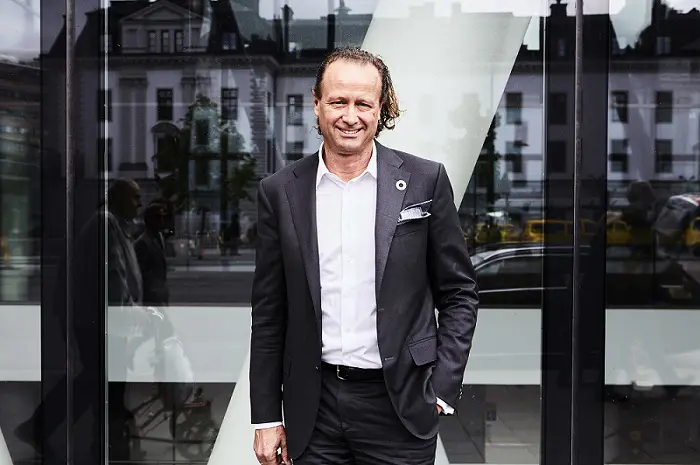Man i kostym står framför en glasbyggnad i stadsmiljö, leende mot kameran