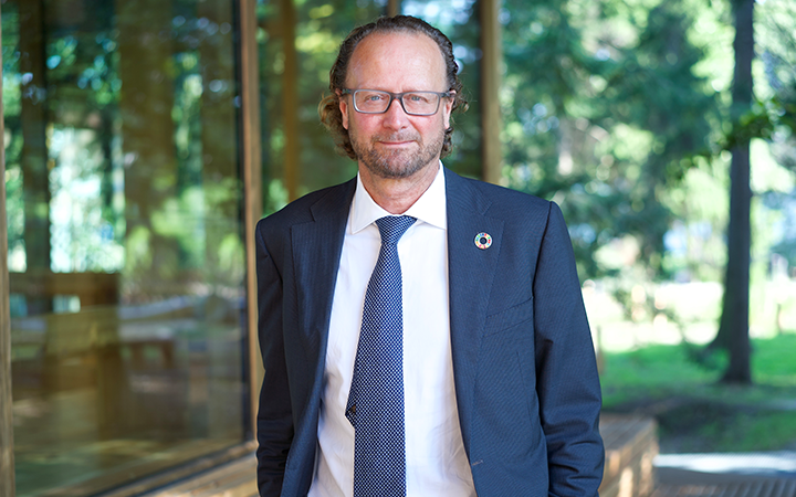 Jan-Erik Saugestad i kostym och slips står utomhus framför en glasvägg, omgiven av träd och grönska