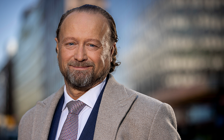 Äldre man i kostym och slips med skägg utomhus mot oskarp stadsmiljö i bakgrunden