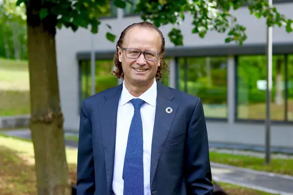 Jan Erik Saugestad i kostym och slips står utomhus framför en modern byggnad med fönster, omgiven av grönska. Han bär glasögon och har långt hår