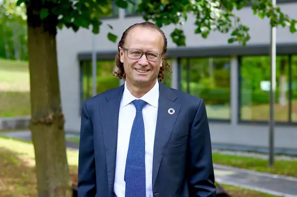 Jan Erik Saugestad i kostym och slips står utomhus framför en modern byggnad med fönster, omgiven av grönska. Han bär glasögon och har långt hår