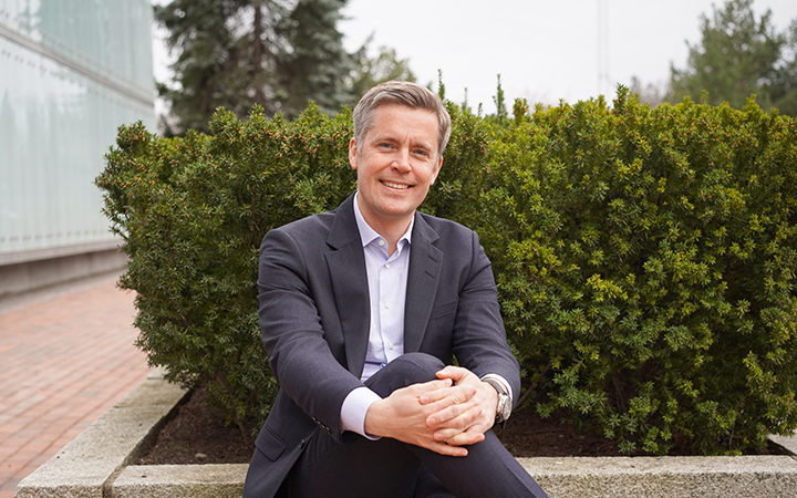 Man i kostym sitter framför en grön buske, leende och ser avslappnad ut utomhus