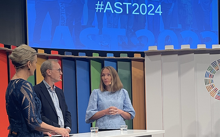 Tre personer diskuterar på en scen under AST2024-konferensen, med en färgglad bakgrund och projektor som visar hashtaggen #AST2024