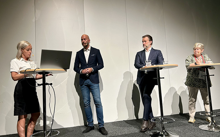 Fyra personer deltar i en paneldiskussion på en scen. En kvinna står vid en dator och talar. Tre män står vid höga bord, två lyssnar uppmärksamt och en ser tankfull ut
