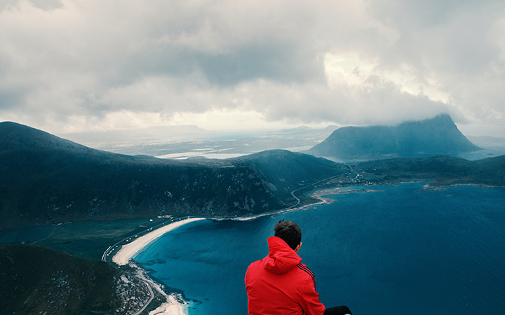 Person i röd jacka sitter på berg och blickar ut över en blå fjord och dramatiskt molnig himmel