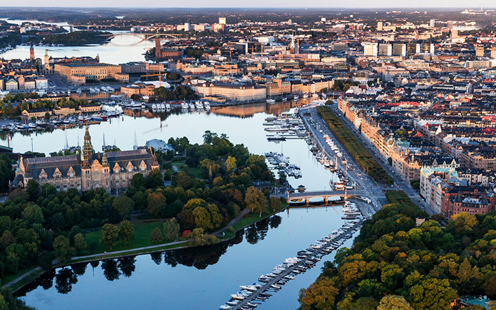 Flygfoto över centrala Stockholm med vy över Djurgården, båtar vid kajen och stadens silhuett i bakgrunden
