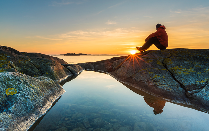 Person i röd jacka sitter på klippa vid solnedgång, speglas i havet, svensk skärgård