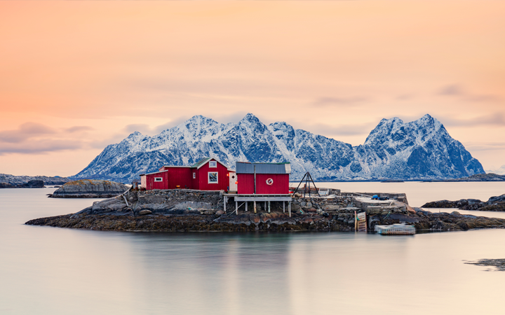 Röda fiskehus på en liten ö vid Lofoten, med snötäckta berg i bakgrunden och en orangefärgad himmel i solnedgången
