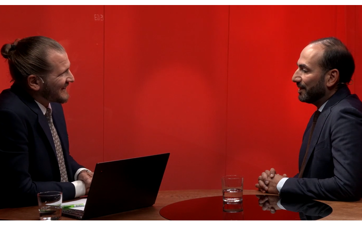 Två affärsmän i kostymer har ett möte vid ett runt bord med en bärbar dator, mot en röd bakgrund