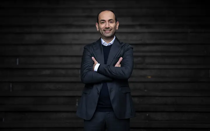 Nader Hakimi i kostym står leende framför trappa, korslagda armar. Mörk bakgrund