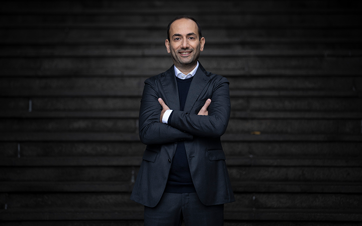 Nader Hakimi i kostym står leende framför trappa, korslagda armar. Mörk bakgrund