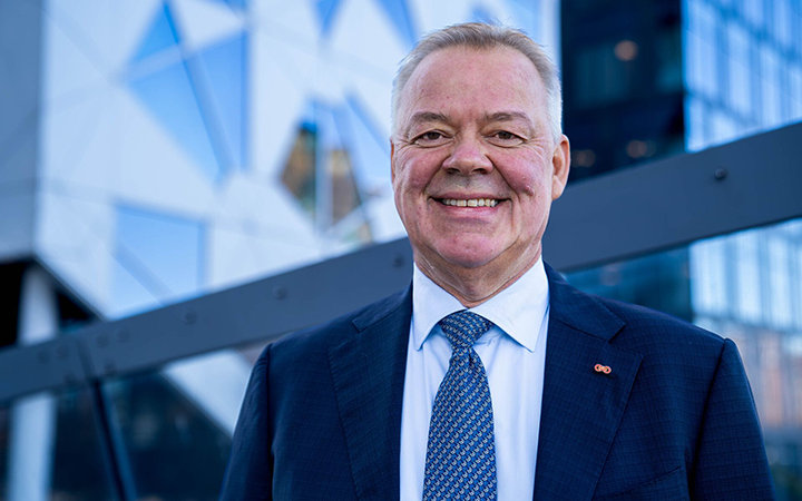 Äldre man i kostym står framför en modern kontorsbyggnad, ler mot kameran