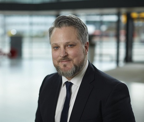 Philip Ripman i kostym med skägg och kort grått hår står inomhus, bakgrund med glasväggar och utsikt