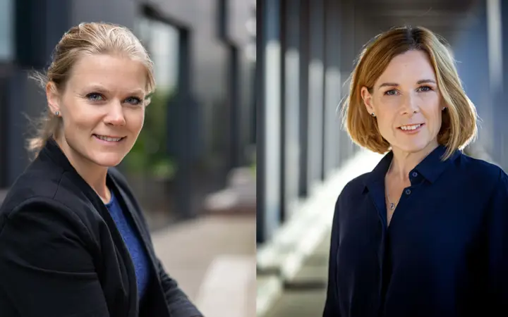 Två kvinnor i affärsmiljö med modern kontorsarkitektur i bakgrunden. De har på sig professionella kläder och ler mot kameran