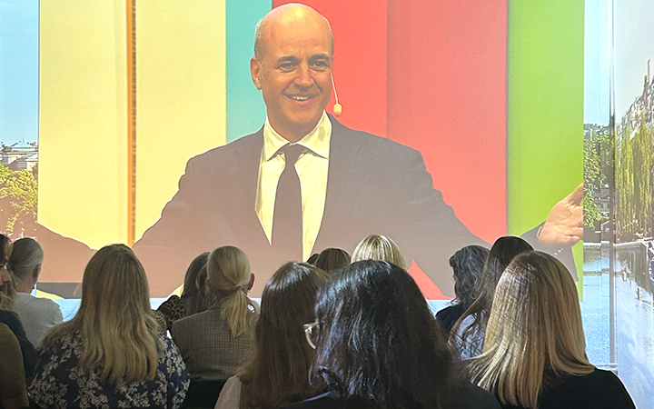 Talare på scenen framför en stor publik under en presentation, med färgglad bakgrund